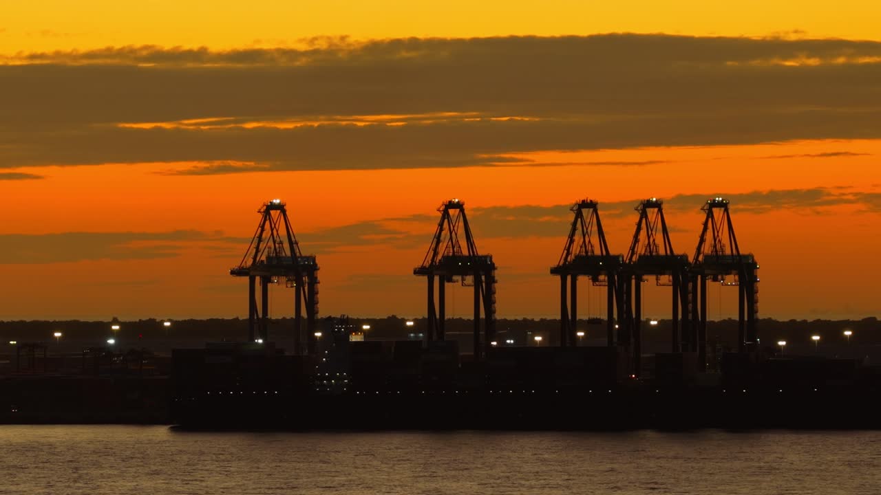Ripresa cinematografica di gru portuali a Puerto Caucedo, in controluce contro un vivace tramonto arancione. Ideale per concetti di commercio globale, logistica e industriali nei Caraibi, Repubblica Dominicana.