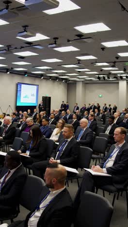 Diverse professionals gathering around conference table, focusing on presenter delivering insights via large projection screen during corporate strategy session