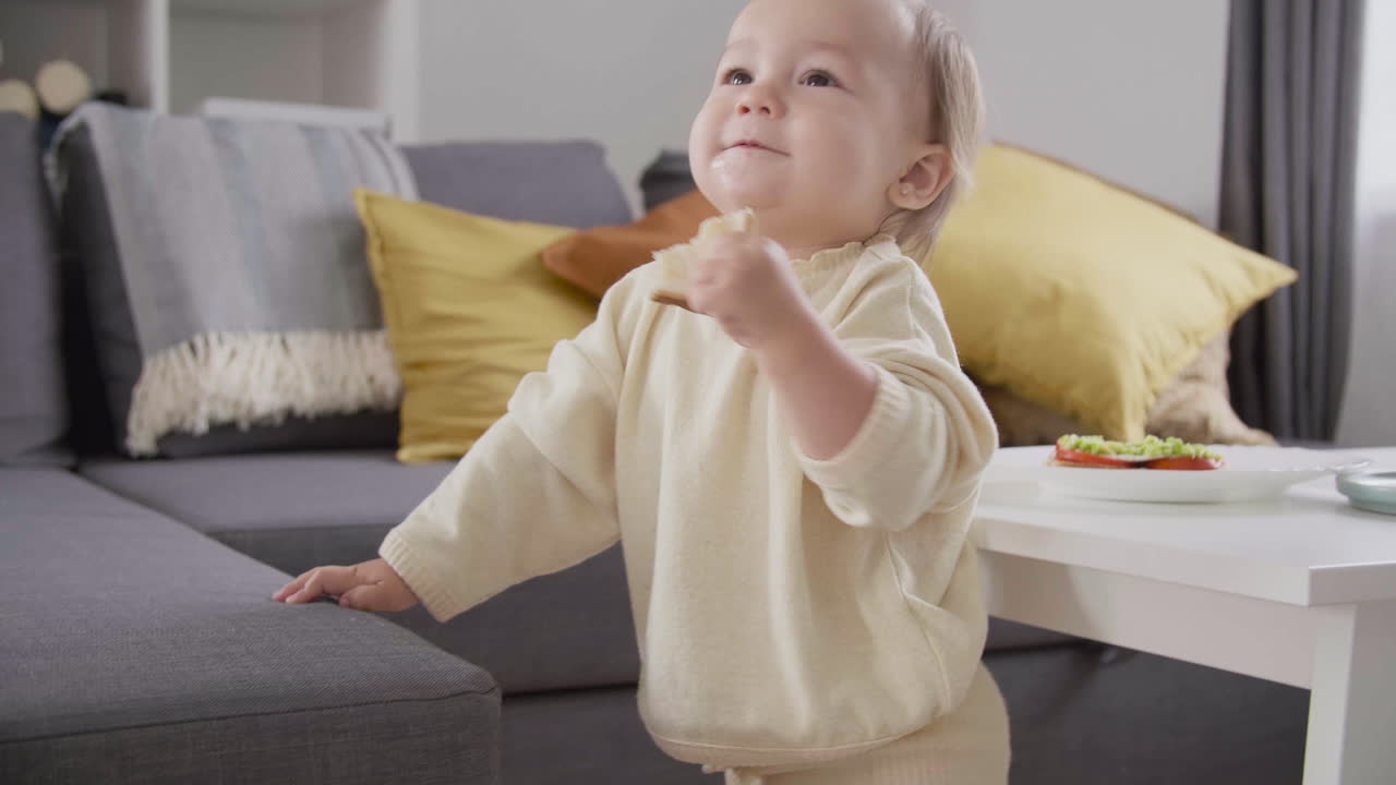 linda niña comiendo pan mientras está de pie junto al sofá en la sala de estar 1