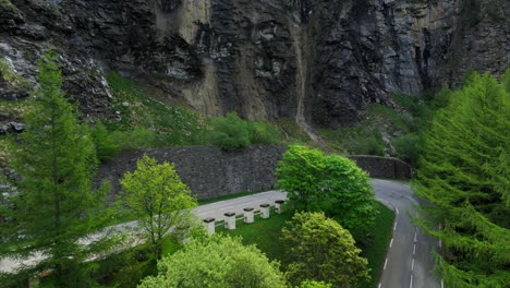 Aerial-view-of-a-black-car-driving-past-a-winding-road-through-Mont-Cenis-with-lush-green-surroundings-and-rocky-cliffs