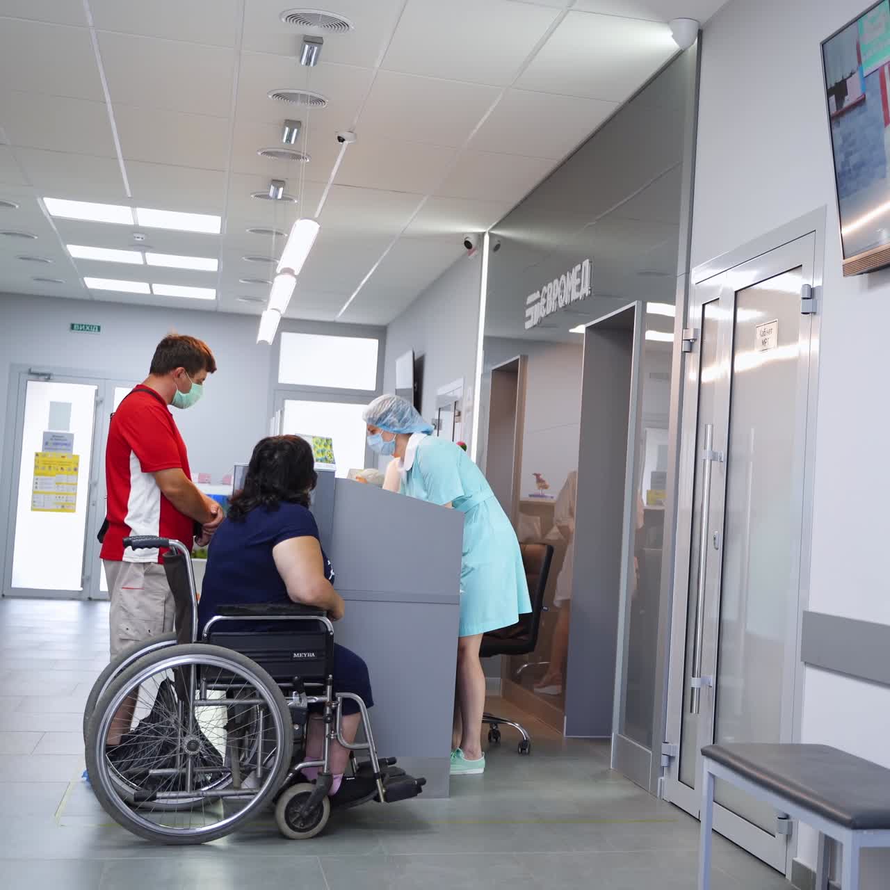 People waiting at the reception in modern hospital. Woman sitting in the wheelchair and man standing beside her addressing the nurse at stand