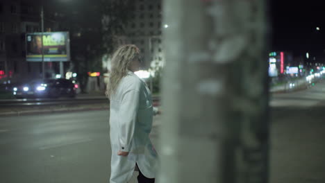 excited young woman in white coat joyfully walks and spins around city street at night, smiling as she plays in open space with glowing car headlights and softly blurred buildings in background