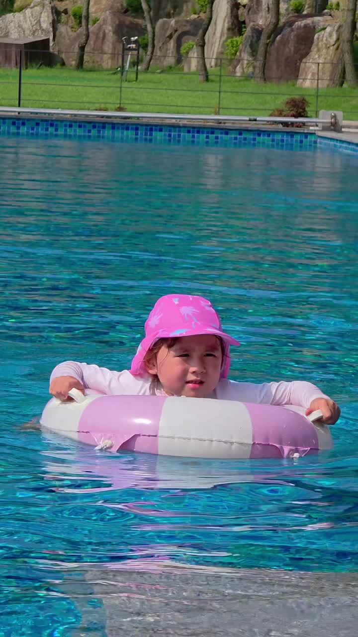 Vertical tracking shot of a happy baby girl in a pink hat learning to swim with an inflatable ring in a beautiful outdoor pool on a sunny summer day