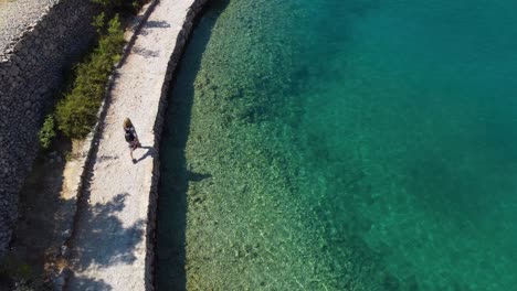 A-mesmerizing-bird's-eye-view-captures-a-blond-girl-walking-on-a-gravel-road-in-Zavratnica-Inlet,-Croatia-and-the-stunning-Mediterranean-coastline-on-a-sunny-summer-day