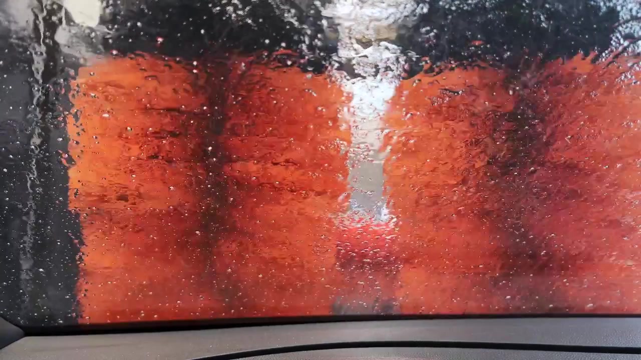 Vista dall'interno di un'auto mentre spazzole arancioni vivaci ruotano contro un parabrezza bagnato in un autolavaggio. Utile per contenuti del settore automobilistico o della pulizia.