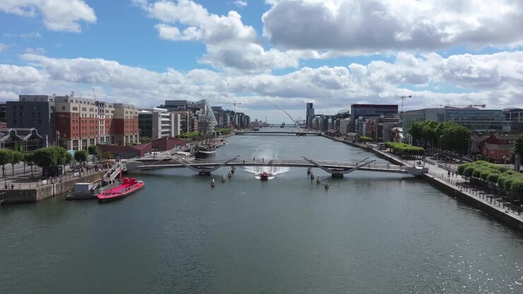 Auto e autobus attraversano un ponte nel centro di Dublino mentre un drone scivola lungo il fiume Liffey, superando moderni uffici e percorsi lungo la riva.
