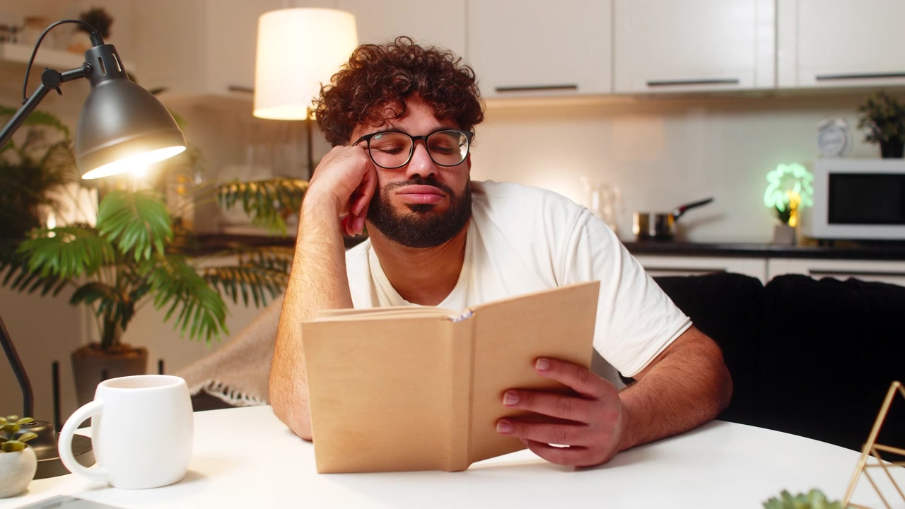 Arabian man reads dull book at night leans on hand bored and tired at desk quiet evening workspace