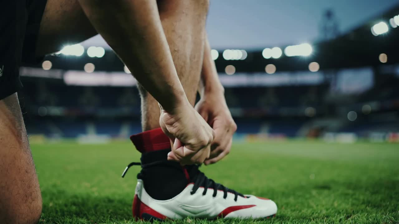 Atleta de camisa vermelha se preparando, amarrando chuteiras vermelhas e brancas em campo de grama e respirando, seguindo o prompt da partida
