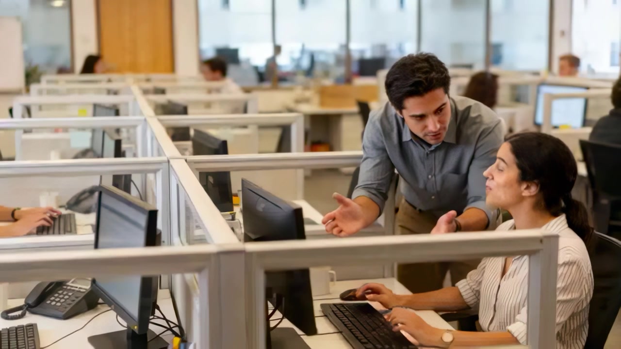 Approaching man leaning explaining screen to seated woman typing at office desk, blouse to assist