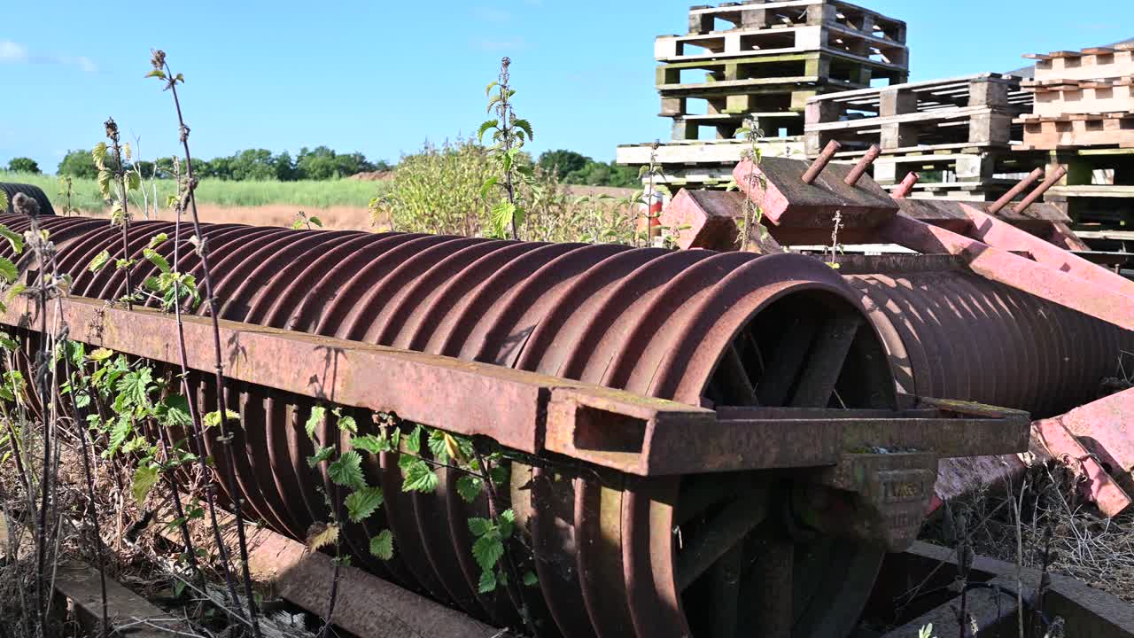 equipos agrícolas viejos cubiertos y abandonados en un patio de la granja de derecha a izquierda con edificios agrícolas y paletas en el fondo