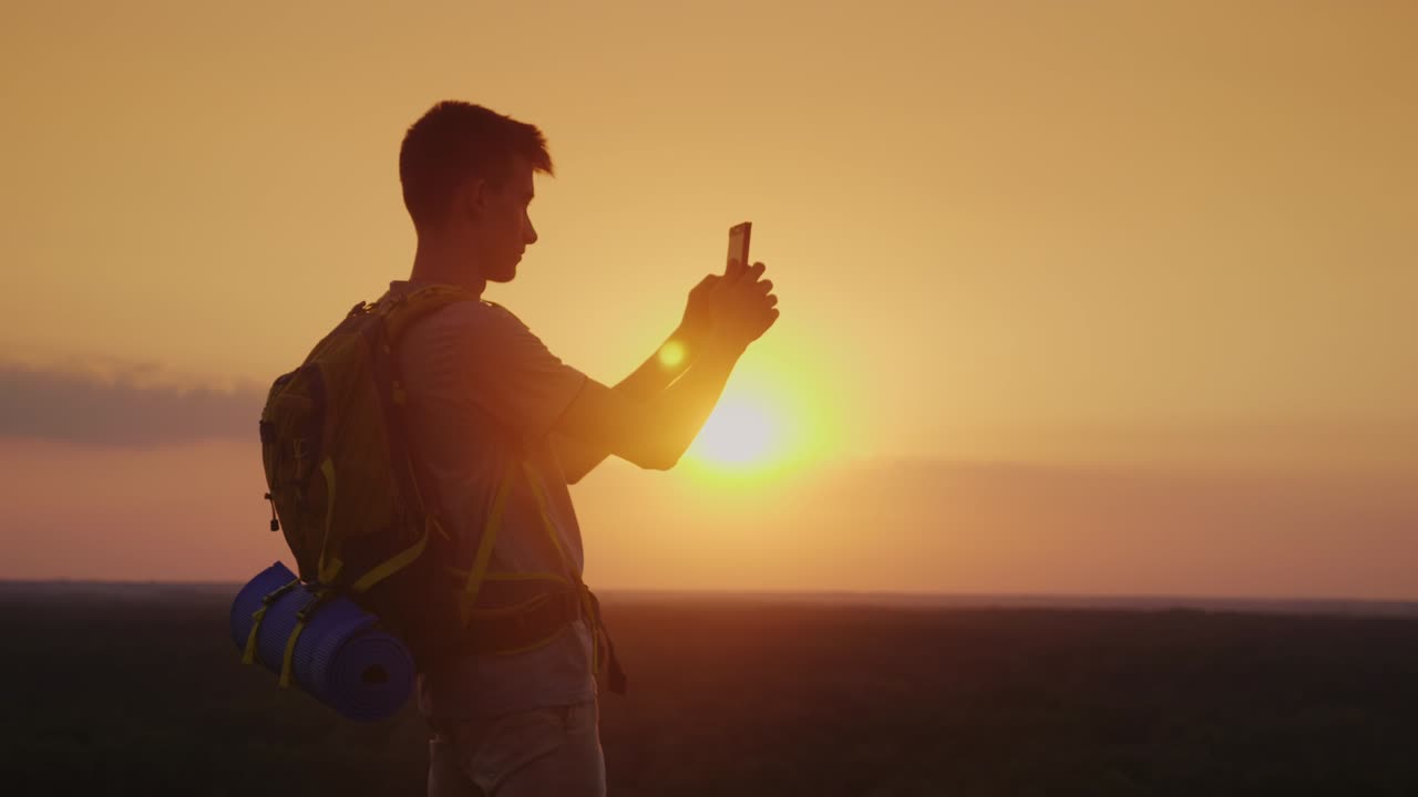 Un giovane con uno zaino sta scattando foto a un bellissimo paesaggio al tramonto. Silhouette su sfondo di cielo arancione.