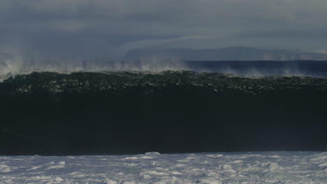 ocean wave sparkles as it catches sunlight glimmer crashing into huge barrel with snow capped mountains behind, slow motion