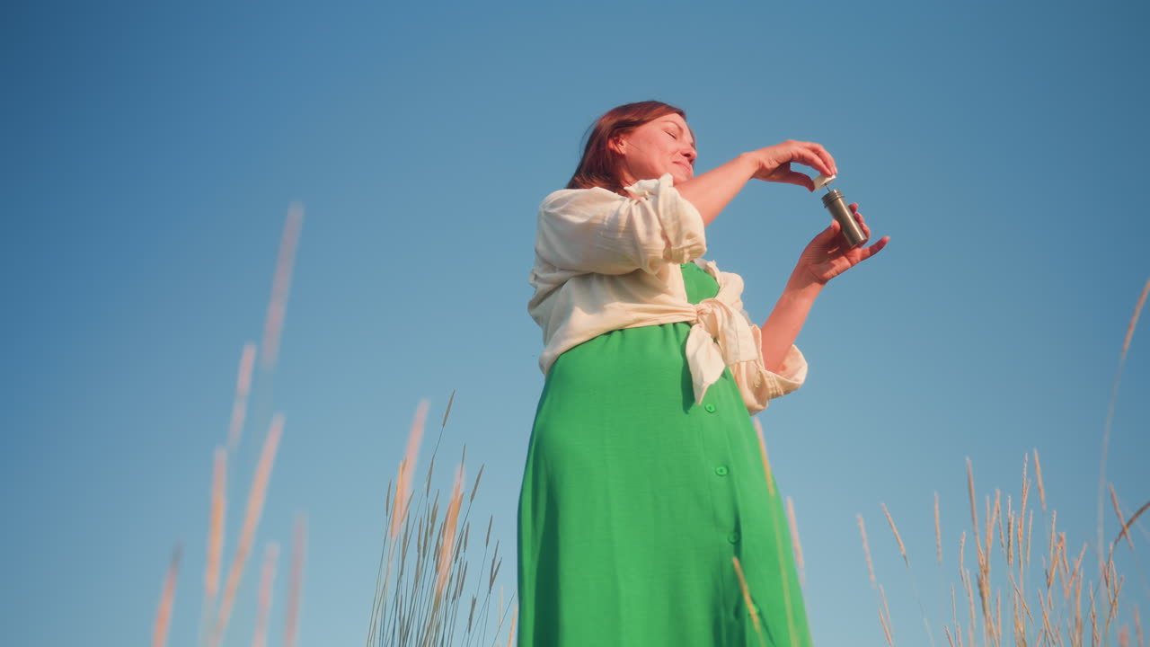 Upward view of woman in green dress standing on grassy hill under clear blue sky blowing iridescent soap bubbles into warm golden hour breeze, tall grass framing silhouette