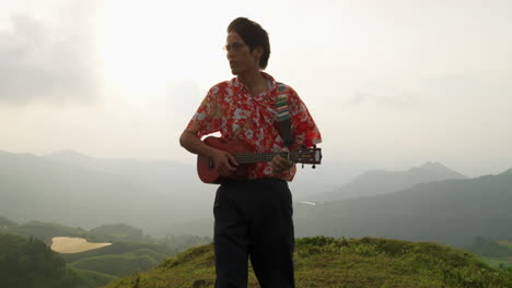 Close-up-shot-of-an-Asian-man-standing-on-top-on-a-mountain-singing-and-playing-ukulele-on-a-cloudy-day