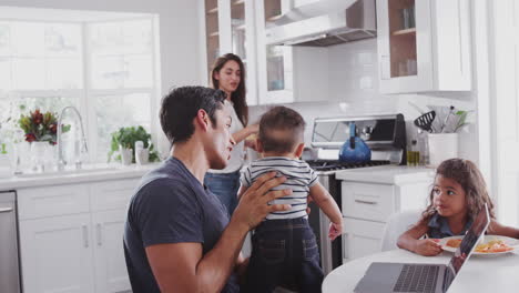 Young-Hispanic-family-in-their-kitchen,-mum-cooking-at-hob,-dad-lifting-baby-in-the-air,-close-up