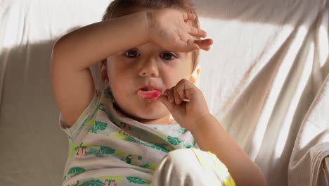 niño pequeño caucásico sentado en una silla vieja y comiendo una piruleta rosa y cubriendo sus ojos