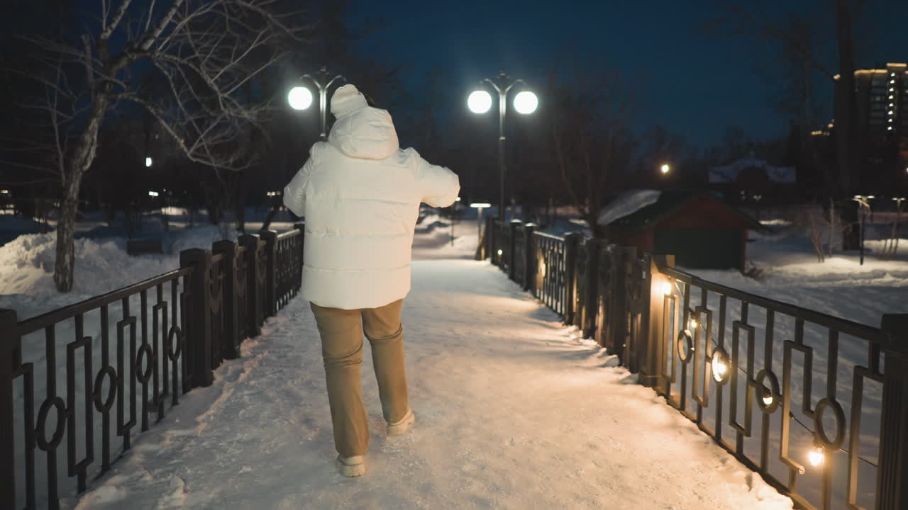 Student wearing white puffer coat and headphones sways along snow covered walkway lined with glowing lights and lampposts under night sky, enjoying rhythmic motion and joyful winter ambiance
