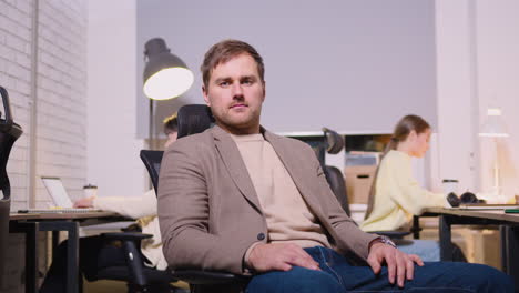 portrait of a confident businessman sitting on office chair and looking at camera while two employees working behind him