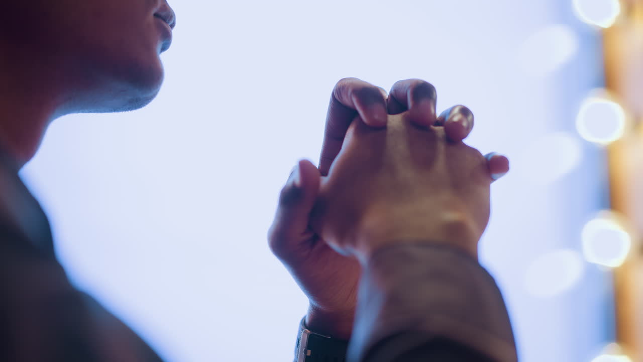 Close-up rear view of young man with clasped hands raised in front of softly lit window, creating mood of calm reflection, quiet emotion, and peaceful thought in ambient indoor setting with warm bokeh lights