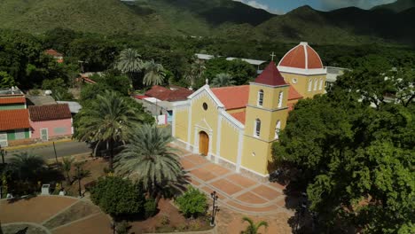 Aerial-reveal-of-San-Juan-Church-in-Isla-de-Margarita,-Venezuela,-surrounded-by-green-trees-and-mountains