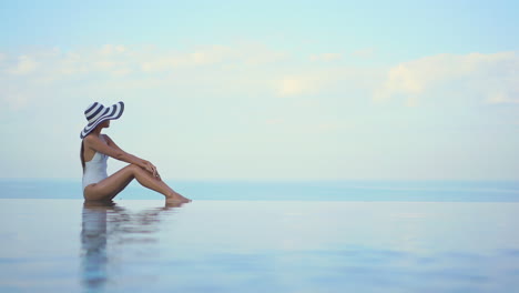 Schöne-Aufnahme-Einer-Eleganten-Asiatischen-Frau-Mit-Großem-Hut-Sitzt-Am-Pool-Und-Sieht-Meer-Aus