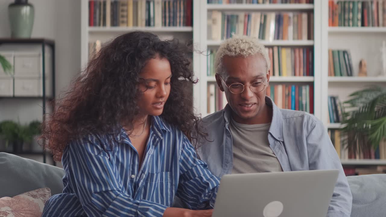 Happy african husband and indian wife relaxing on sofa enjoying using laptop