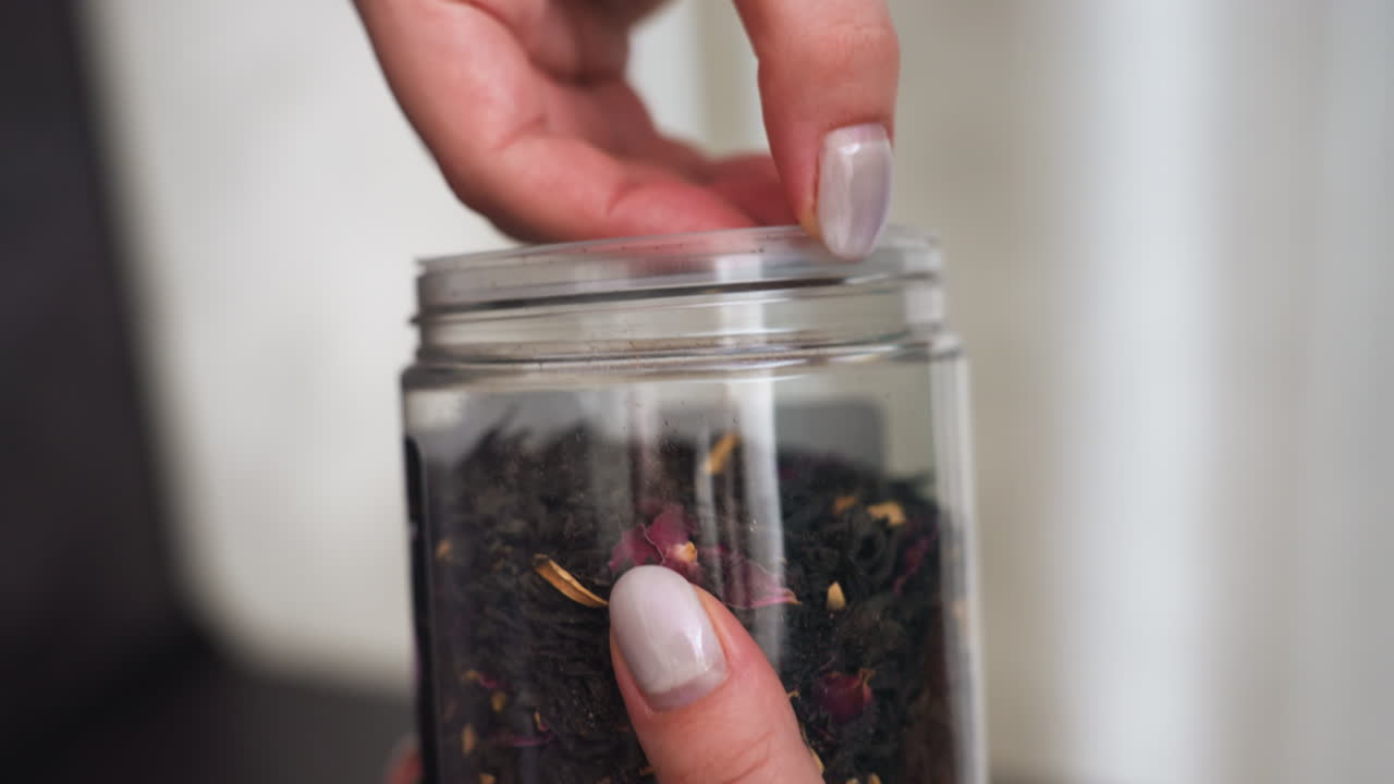 Caucasian Woman Lifting Glass Lid, Woman Carefully Opening Glass Jar With Loose Leaves Inside In Kitchen, Closeup Shot Of Woman Opening Glass Container Filled With Dark Loose Leaves In Warm Kitchen
