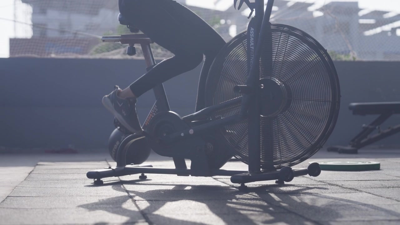 Tracking shot of the lower body of a South Asian female athlete pedaling an assault air bike during functional fitness workout in a modern indoor fitness studio. Collection: WORKOUTSERIES2026.