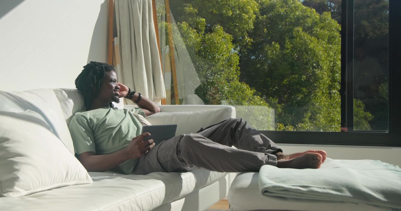 African American man starting with tablet while relaxing on sofa, tapping and glancing out window