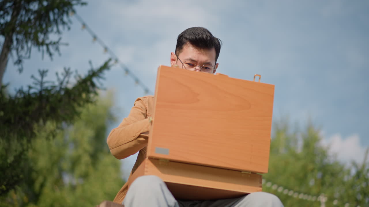 Seorang seniman Asia membuka kotak easel kayu di bangku taman di bawah lampu senar, menyiapkan palet dan kuas untuk melukis di udara terbuka, ekspresi fokus, jaket cokelat kasual, pohon pinus, dan latar belakang langit biru
