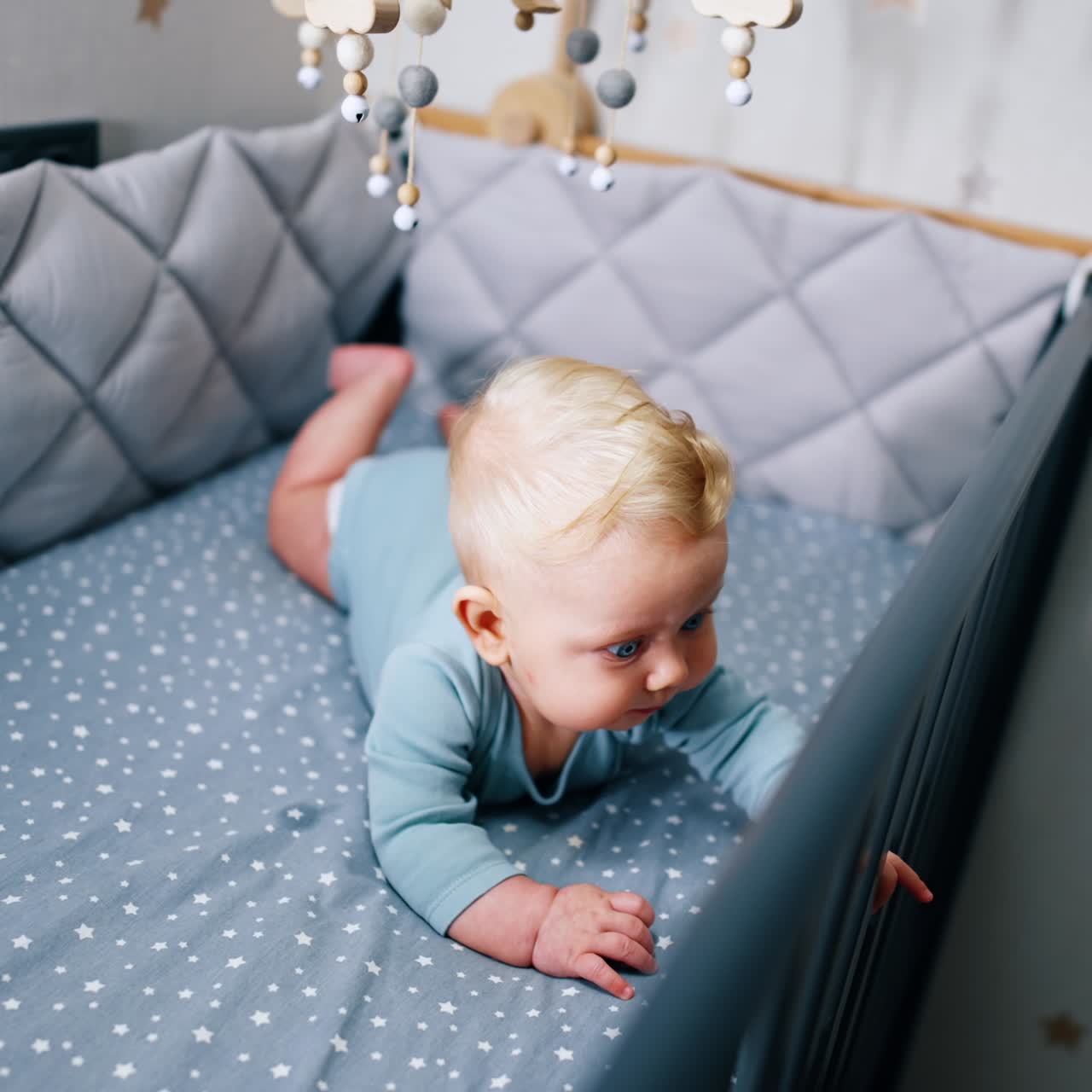Little cute child lying on the belly. Adorable infant baby in bodysuit lies in cot under the crib mobile