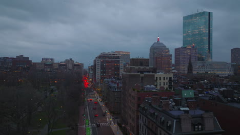 Evening-flight-above-park-and-buildings-in-urban-borough.-Traffic-on-streets.-Modern-high-rise-building-against-sky-at-dusk.-Boston,-USA