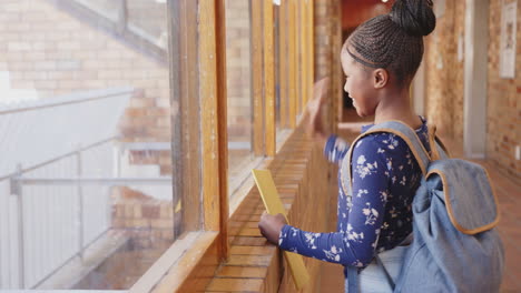 in school, girl with backpack waving through window, holding yellow folder, copy space
