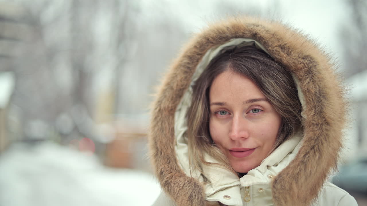 A woman stands in a snowy area, wearing a cozy coat with a fur hood. She displays a calm expression while surrounded by winter scenery and falling snowflakes