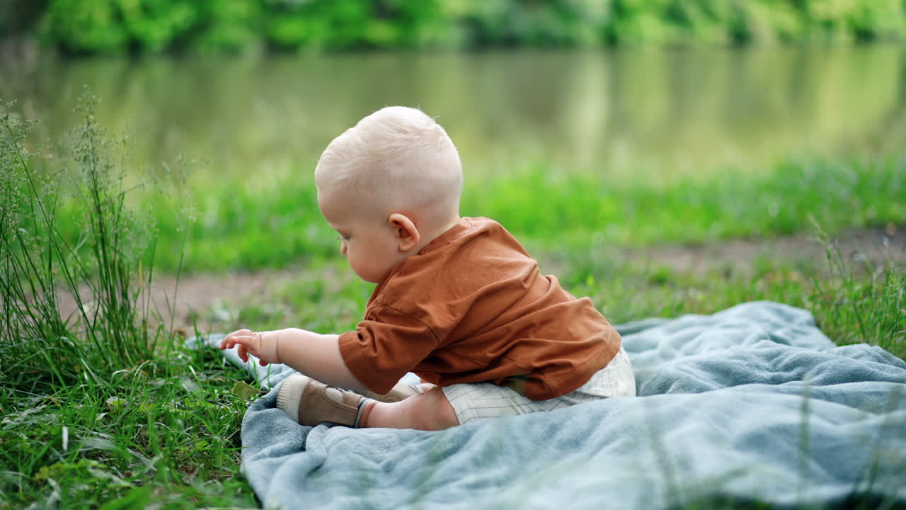 Adorable baby boy sits on the plaid in the grass. Toddler kid touches the grass with interest. Blurred backdrop.