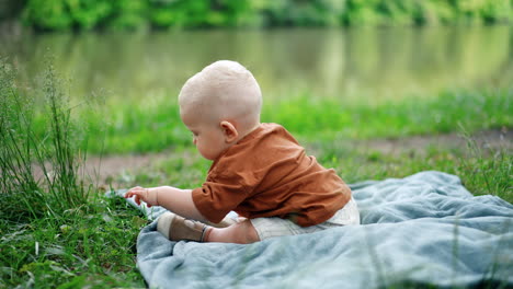 adorable baby boy sits on the plaid in the grass. toddler kid touches the grass with interest. blurred backdrop.