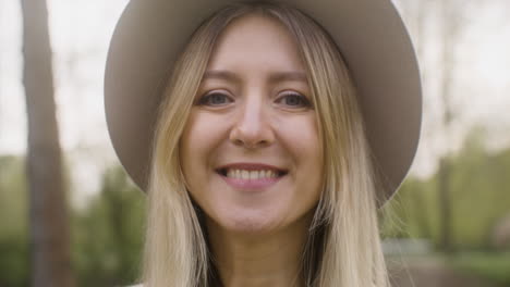 retrato de una mujer rubia feliz con sombrero de pie en el parque y mirando a la cámara 1