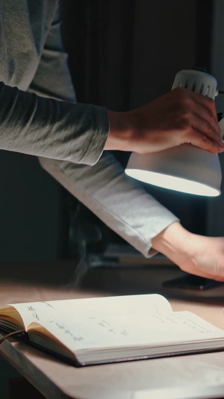 Vídeo vertical: Braços alcançando, adulto com blusa cinza inclinando luminária na mesa para ler um caderno.