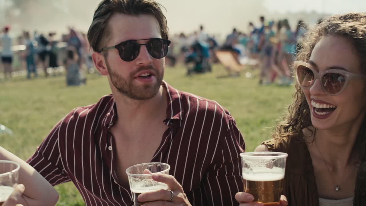 Gruppe af venner sidder sammen på græsset til en musikfestival og drikker øl, mens de snakker.