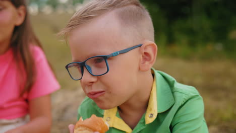 niño pequeño con camisa verde y gafas disfruta de un rollo de salchichas mientras está sentado al aire libre con su familia, el fondo presenta campos abiertos y balas de heno, capturando momentos pacíficos y alegres de comidas al aire libre