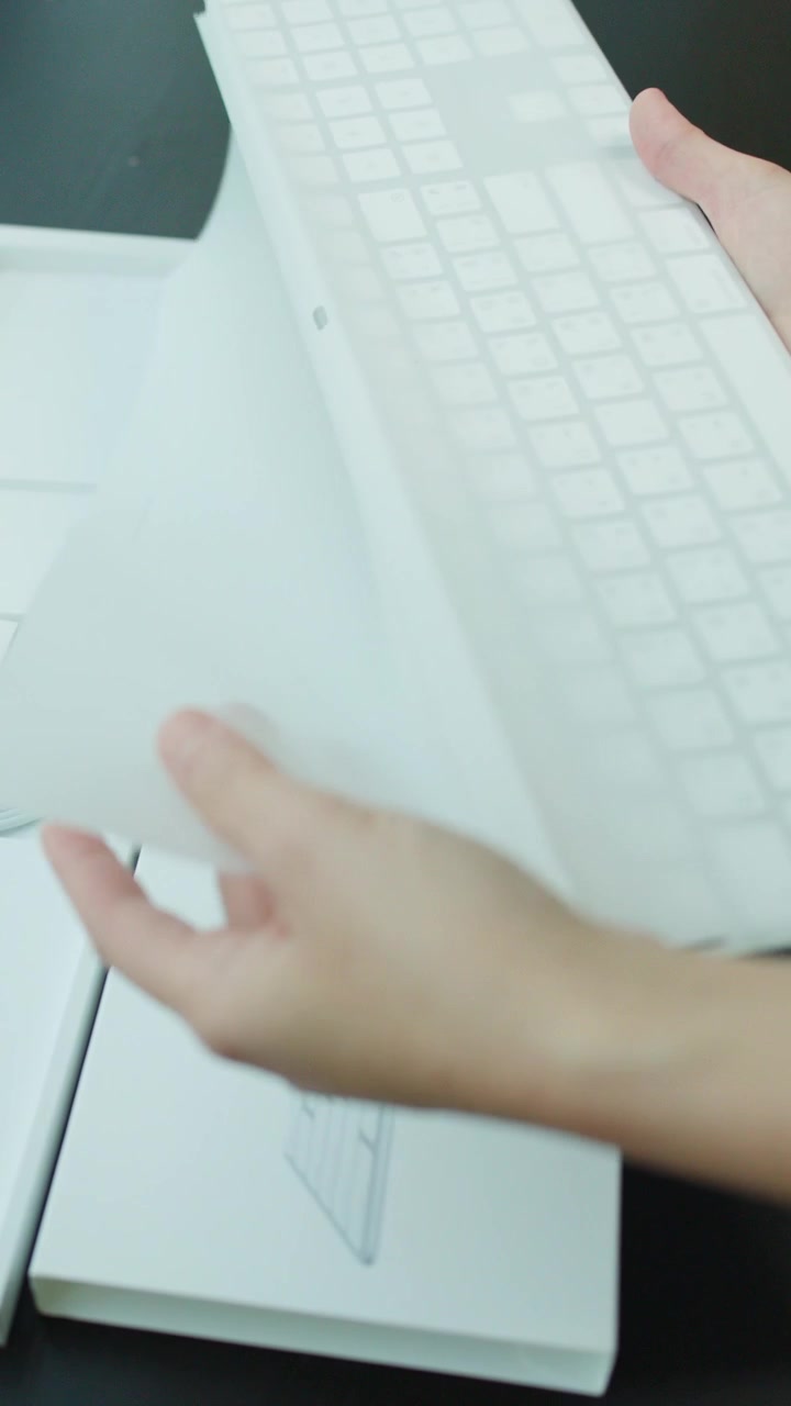 Hands unbox, unwrap, and test a wireless keyboard on a dark desk with soft lighting
