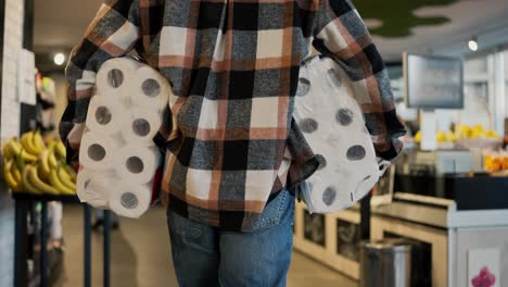 Rear-view-of-a-man-in-a-plaid-shirt-and-blue-jeans-carrying-two-large-packages-of-white-disposable-towels-while-walking-near-the-shelves-in-a-modern-grocery-store