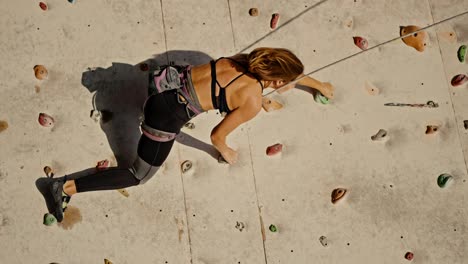 Vertical-video-of-a-blonde-girl-with-a-bob-hairstyle-in-a-black-top-and-a-special-uniform-and-equipment-for-folding-using-belay-climbing-up-the-route-of-a-climbing-wall-in-the-summer