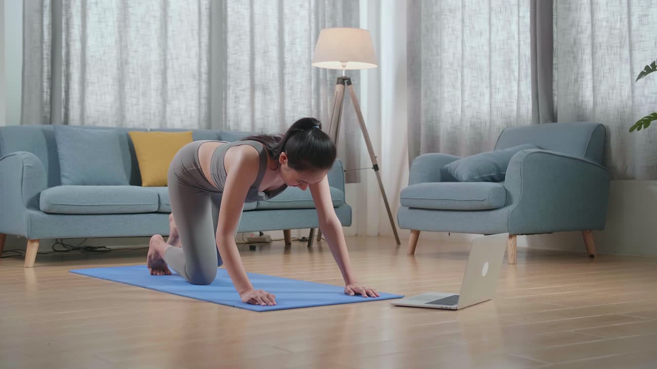 スポーツ服を着た若いアジア人アスリートの女性がラップトップコンピュータを通じてオンラインコーチと一緒に家でマットの上でバランステーブルポーズでヨガをしています