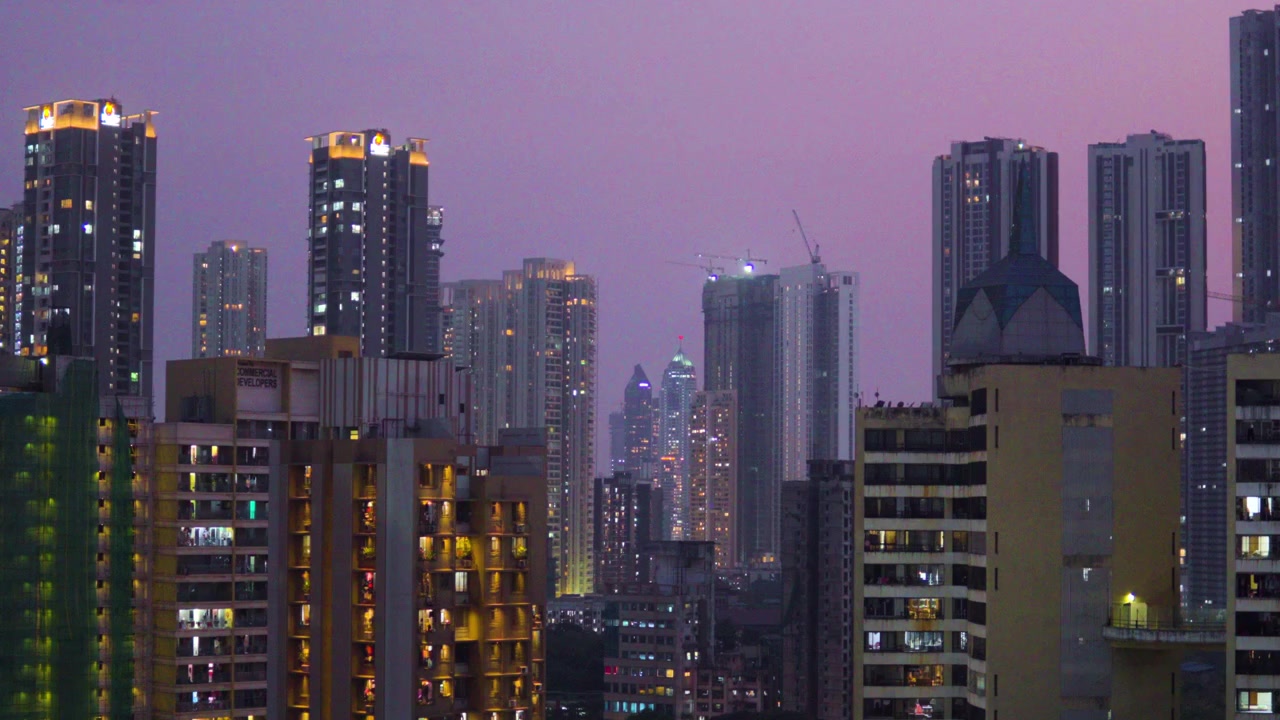 Uma ampla tomada cinematográfica do horizonte da cidade de Mumbai ao anoitecer, com arranha-céus iluminados, guindastes de construção, tráfego em movimento e uma roda-gigante brilhante sob um céu crepuscular roxo.