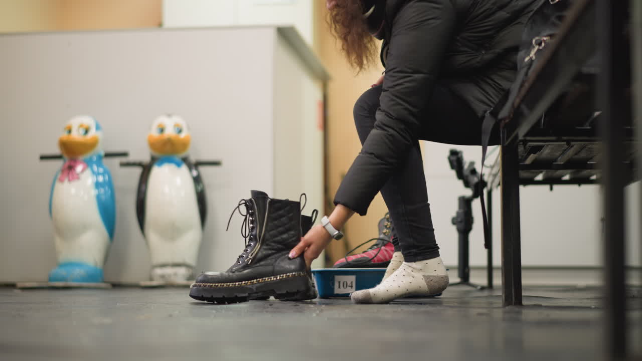 Female tying black leather boots with thick soles and chain detail while sitting on bench wearing polka dot socks, with penguin skating aids visible in background creating indoor winter sports preparation