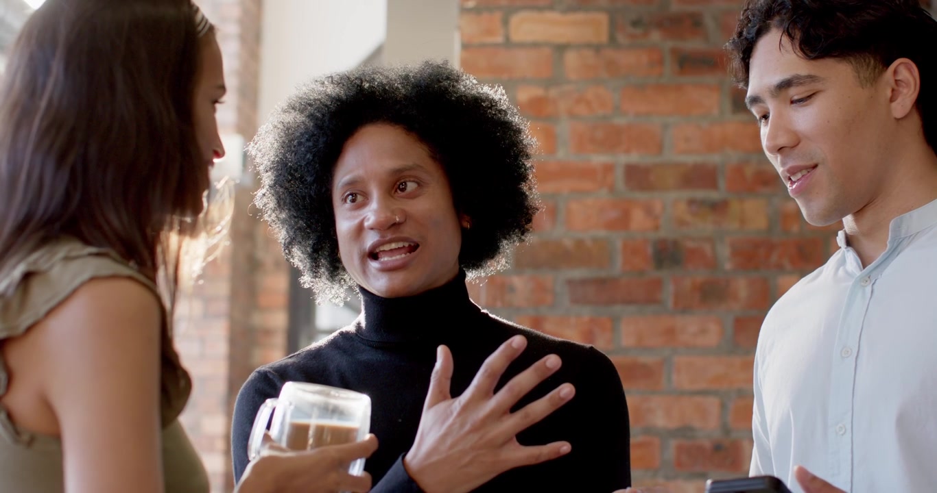 Diverse team in loft listening to lead presenting in turtleneck making eye contact with mug, tablet