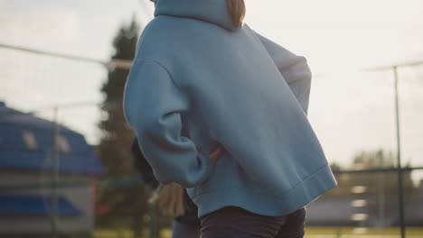 primer plano de una joven con capucha azul realizando ejercicios de fitness al aire libre, la luz del sol brillando por detrás, creando sombras dinámicas y un efecto de iluminación suave en su silueta en la cancha al aire libre