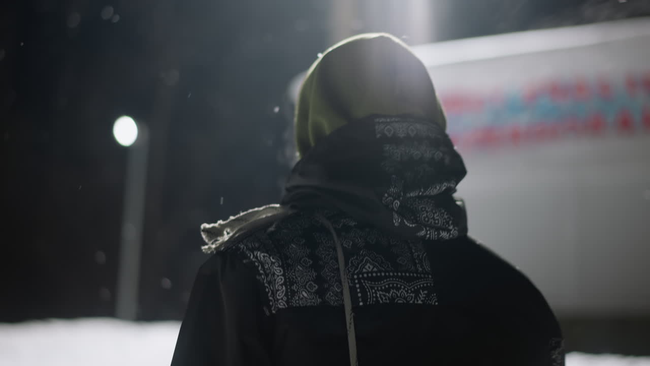 Back view of person walking through snowy area during winter night with visible cold air vapor escaping mouth, wearing hooded jacket under bright artificial lighting and soft focus background