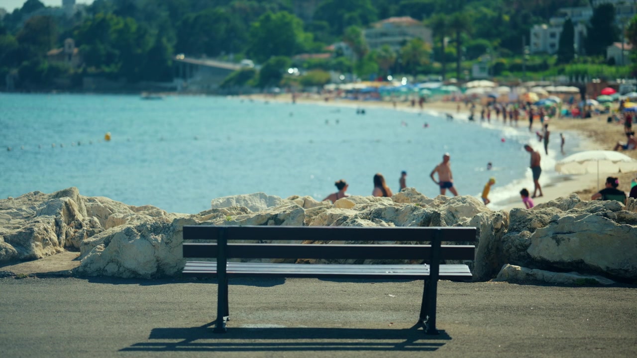 Bænk med udsigt over havet, der tilbyder et afslappende sted at se folk nyde den sandede strand og det rolige vand under sommersolen